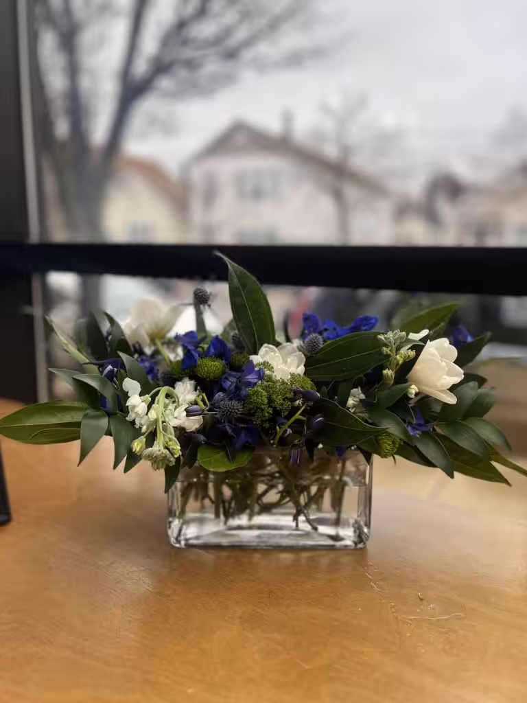A rectangular glass vase holds a bouquet of white and deep blue flowers with green leaves on a wooden table