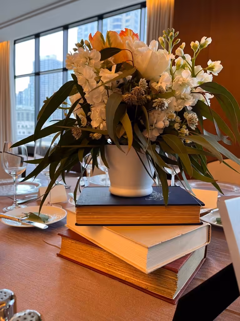 Vase of white and orange flowers sits on a stack of three hardcover books on a dining table
