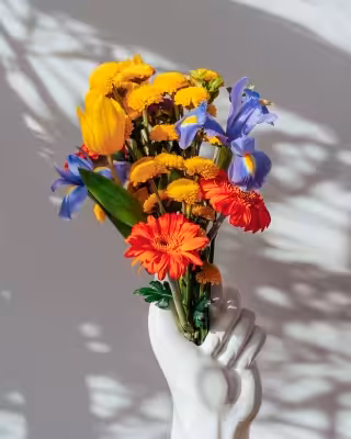 Bouquet with orange, yellow, and purple flowers is held by a white ceramic hand, casting soft shadows on a neutral background